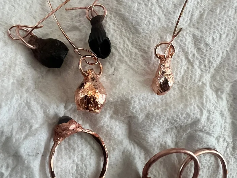 Small copper jewellery pieces and wrapped dark stones arranged on pale textured fabric.